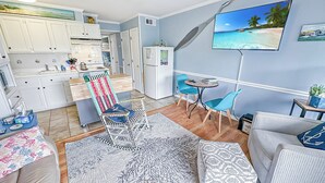 A compact living space with a kitchenette, a dining area with a small table and chairs, a sofa, and a TV mounted on the wall. - “TYBEE ISLAND DREAMING”Private Balcony Overlooking the Ocean, Beautiful View.  (Tybee Island)