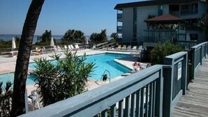A pool area with a view of the ocean, a building with balconies, and a wooden deck. - “TYBEE ISLAND DREAMING”Private Balcony Overlooking the Ocean, Beautiful View.  (Tybee Island)