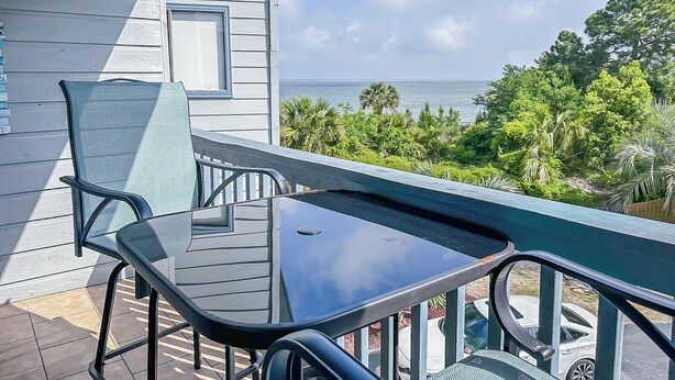 A balcony with a glass table and chairs, overlooking a view of the ocean and lush greenery. - “TYBEE ISLAND DREAMING”Private Balcony Overlooking the Ocean, Beautiful View.  (Tybee Island)