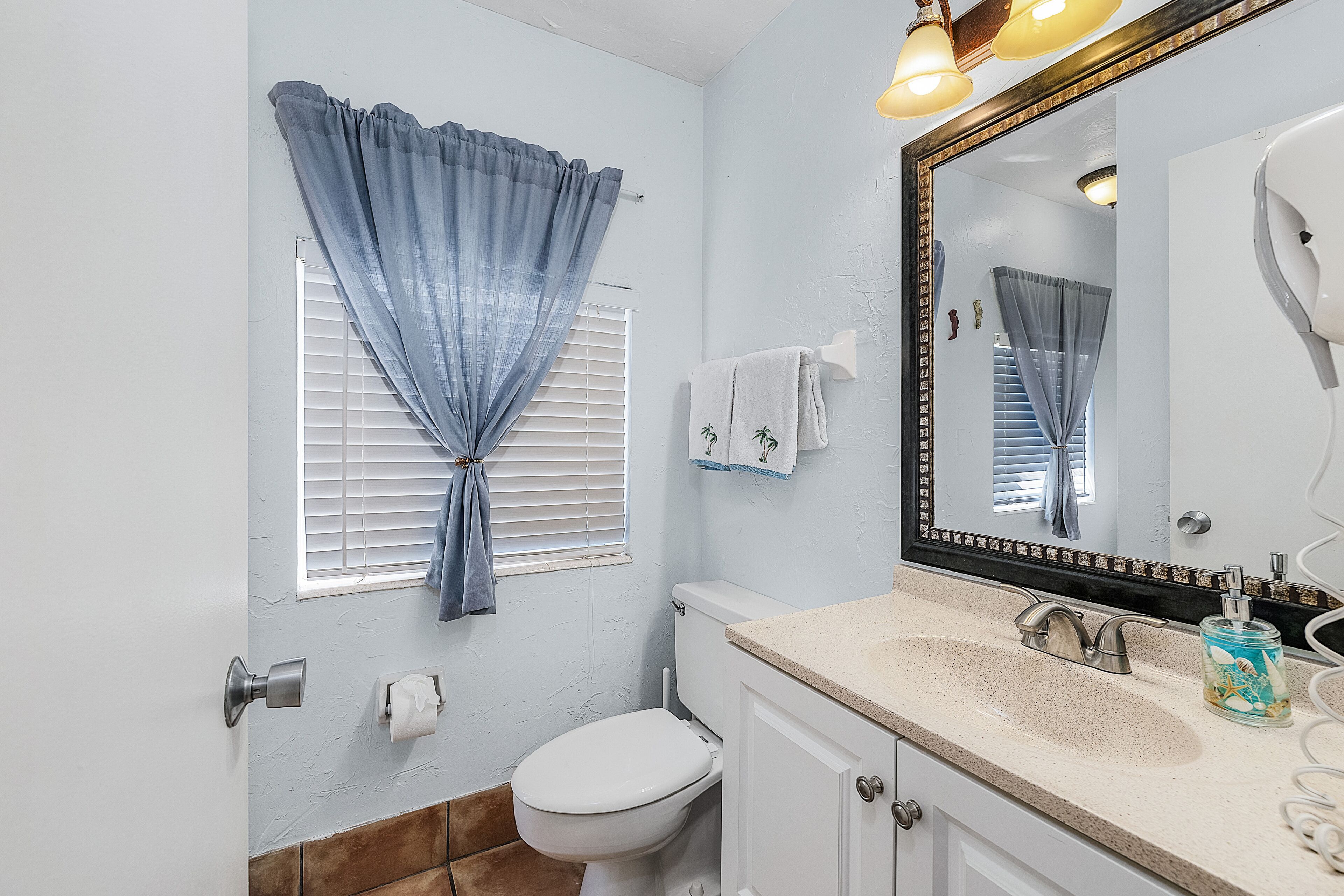 Une salle de bain équipée de toilettes, d’un lavabo, d’un miroir et d’une fenêtre avec des rideaux.
