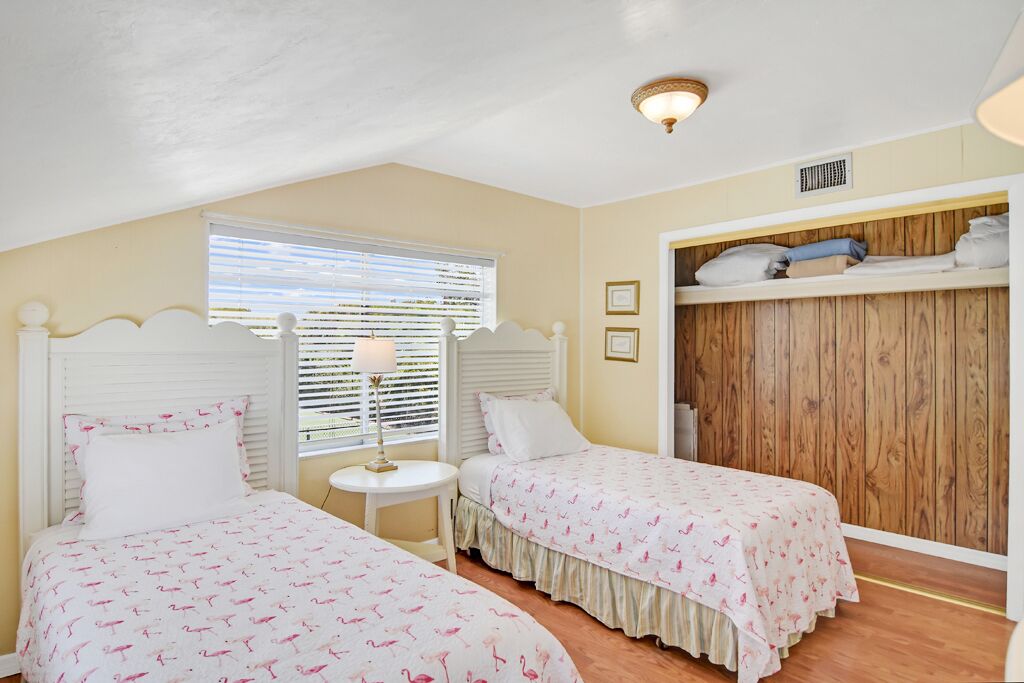 A bedroom with two beds, a window with blinds, a bedside table with a lamp, and a wooden wardrobe.