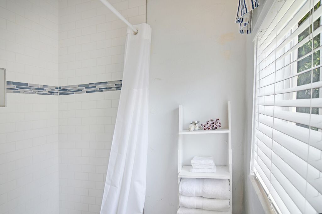 Un baño con paredes de azulejos blancos, cortina de ducha y una ventana con persianas.