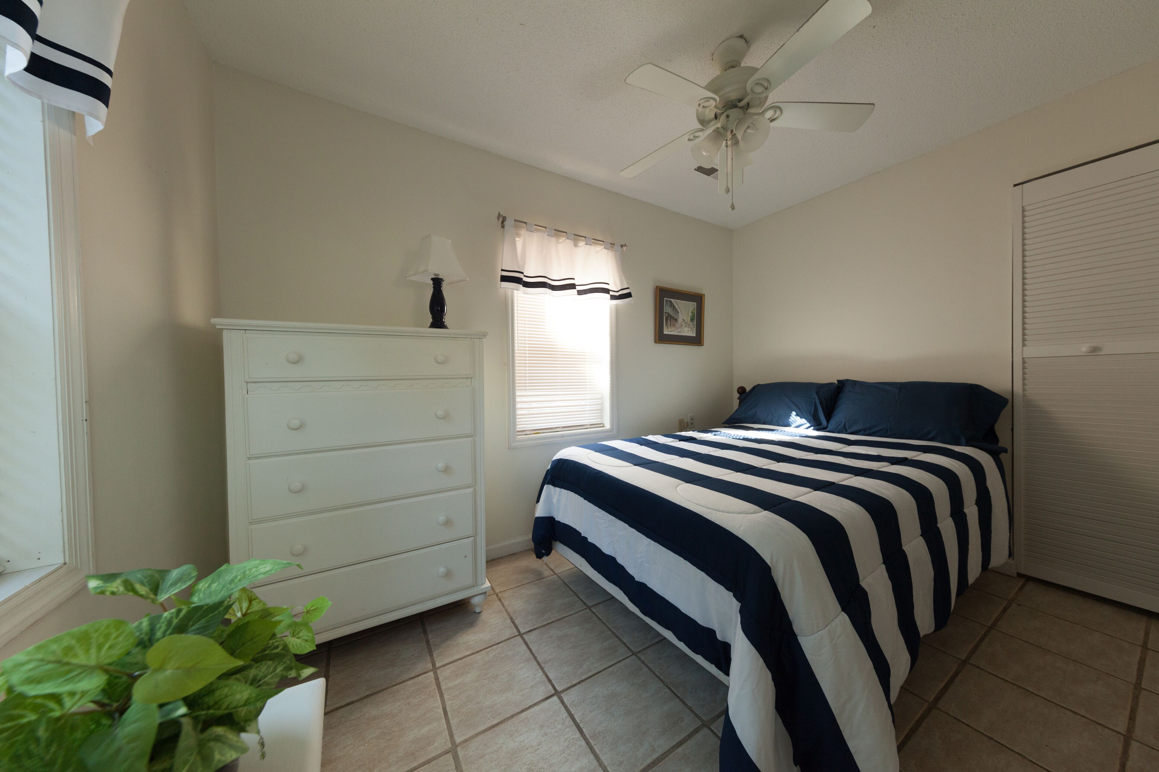 A bedroom with a bed, a dresser, a ceiling fan, a window with blinds, and a potted plant.