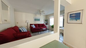 Ein Wohnzimmer mit einem roten Sofa, einem grünen Sofa und einem Couchtisch. Es gibt einen Deckenventilator, eine Lampe und ein gerahmtes Bild an der Wand.
