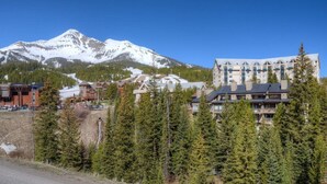 A mountain resort with a prominent snow-capped peak, a large lodge with multiple floors, and dense evergreen trees in the foreground. - Slope-Side 1 Bedroom, Walk to Chairlifts! (Big Sky)