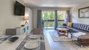 A modern living room with a sofa, a dining table set with plates and glasses, a TV mounted on the wall, and a balcony with patterned curtains. - Slope-Side 1 Bedroom, Walk to Chairlifts! (Big Sky)