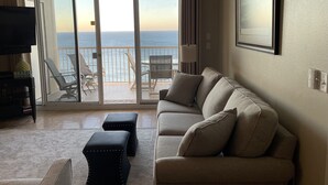 Ein Wohnzimmer mit einer Couch, Hocker, einem Fernseher und einem Balkon mit Meerblick.