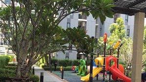 A playground with slides and climbing structures, surrounded by greenery and a building in the background. - ICONDO Ngamwongwan 1B by Tanny (Nonthaburi)