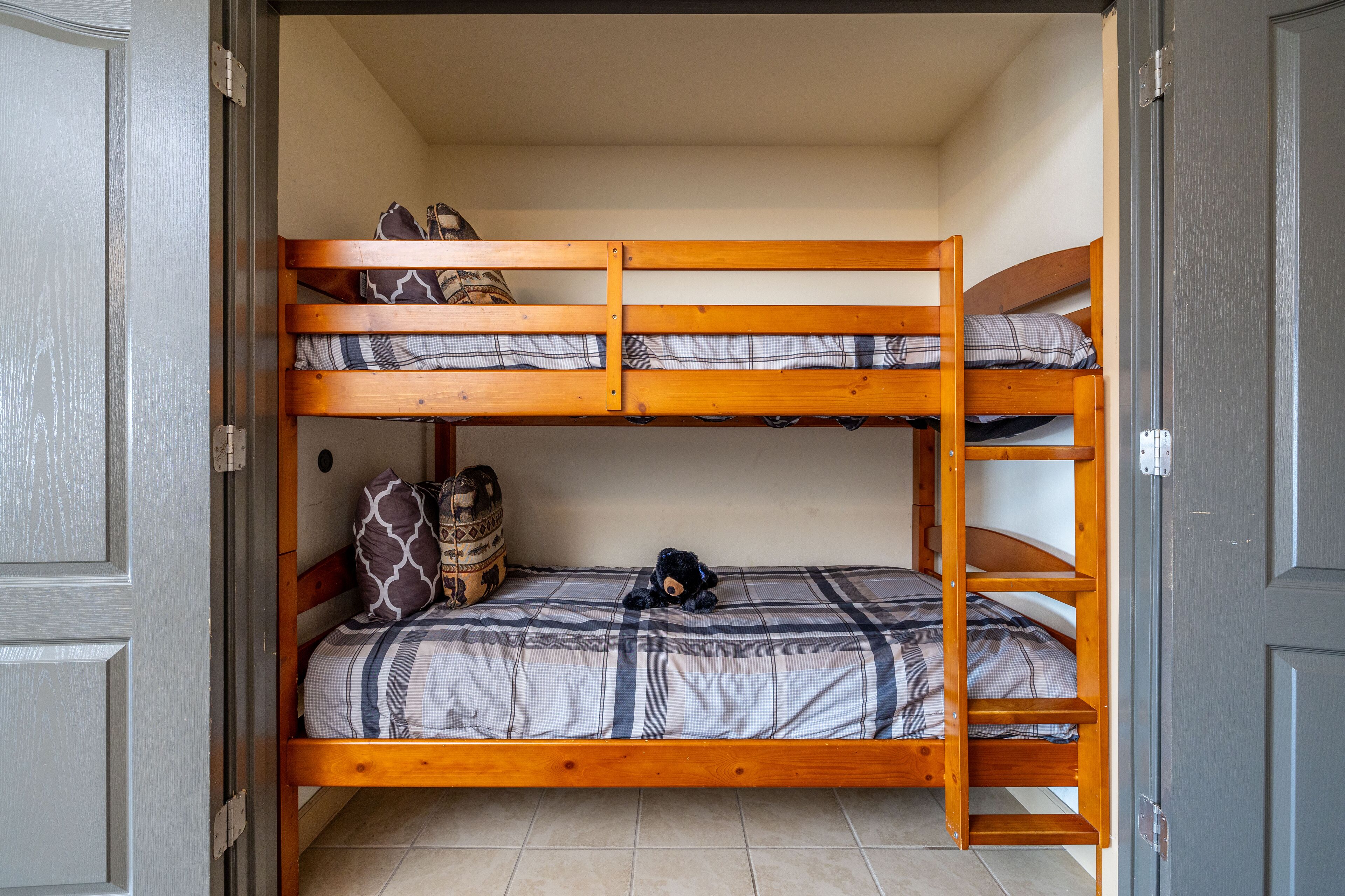 Une chambre avec un lit superposé, une couette à carreaux, un ours en peluche et un sac à dos posé sur le lit du bas.