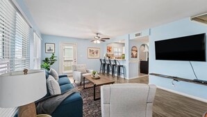 A modern living room with a sofa, armchair, and coffee table. There is a dining area with a table and chairs, and a kitchen in the background. - Beachside home in Sea Isle with sunset & waterfront views, 1-min walk to beach! (Galveston)