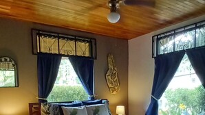 A bedroom with a wooden ceiling, a bed with a floral bedspread, two nightstands with lamps, and windows with curtains. - Unique Jungle Luxury for 5, Fantastic Views, A/C, Jacuzzi Spa & Private Pool! (Arenal Volcano National Park)