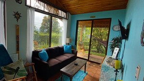 A cozy living room with a brown sofa, blue cushions, a wooden coffee table, and a view of lush greenery outside. - Unique Jungle Luxury for 5, Fantastic Views, A/C, Jacuzzi Spa & Private Pool! (Arenal Volcano National Park)