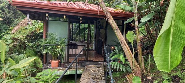 A covered outdoor area with a stone floor, surrounded by lush greenery and potted plants.