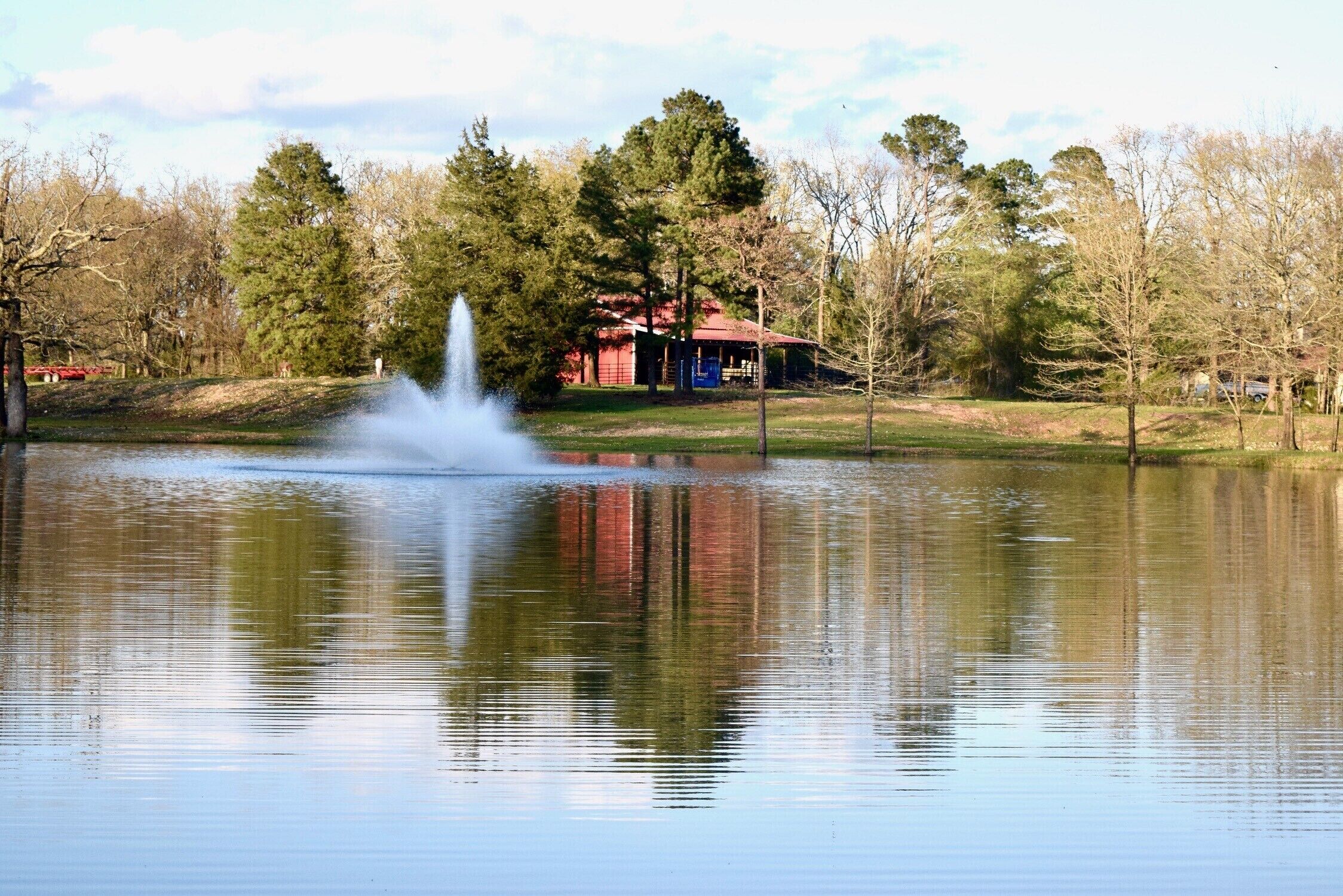 A tranquil lake with a fountain in the center, surrounded by trees and a small building with a red roof.