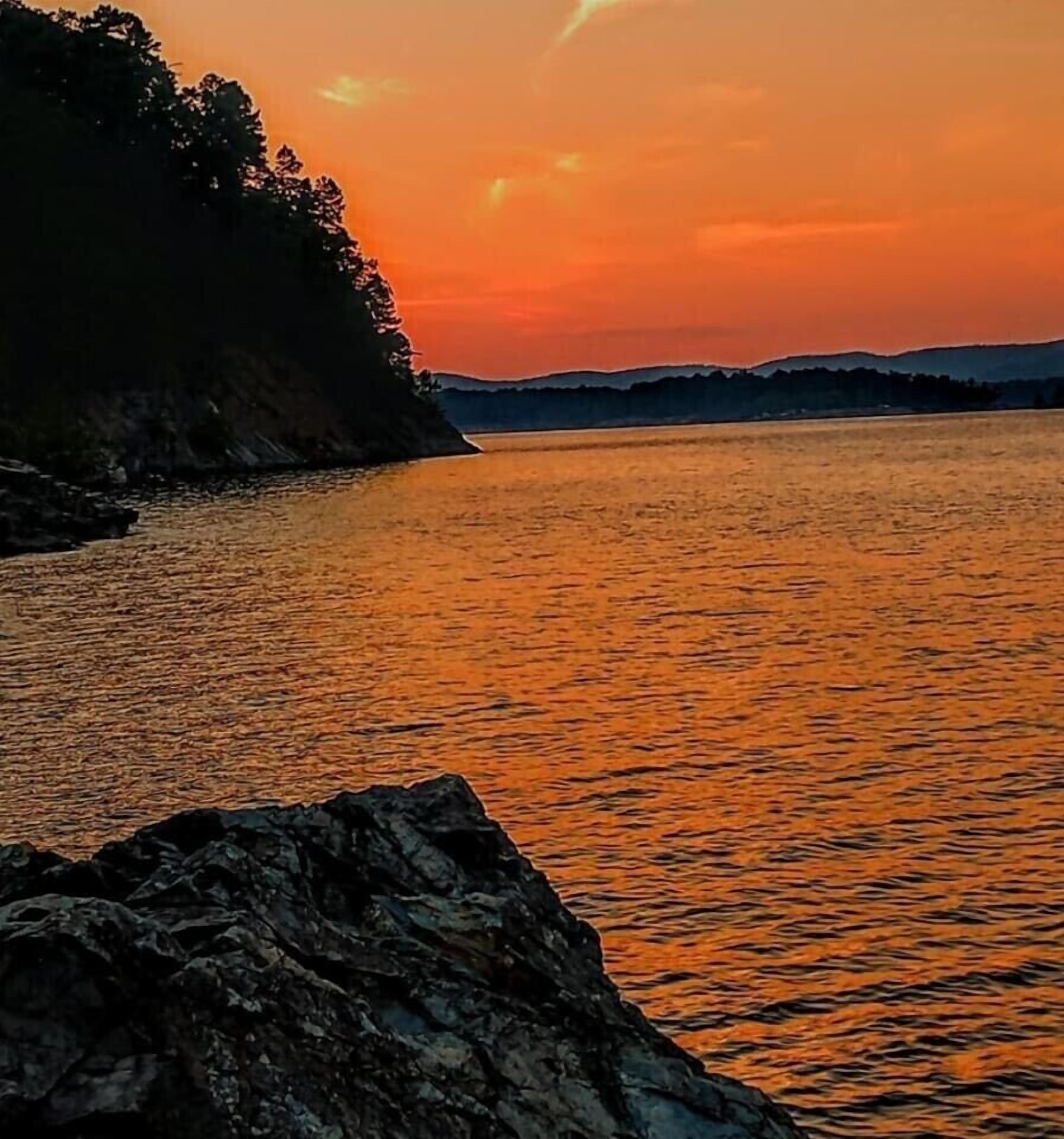 A sunset over a calm body of water with a rocky shoreline and distant hills.