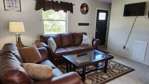 A living room with a brown leather sofa, a glass coffee table, a lamp, a patterned rug, a door, and a wall clock.