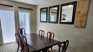 A dining area with a dark wooden table and six chairs, a large window with white curtains, and three framed mirrors on the wall.