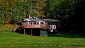 A wooden cabin with a deck and a covered porch, surrounded by a grassy field and a pond. - Cooperstown Dreams Park, All Star Village & Hall of Fame cottage rental. (Milford)