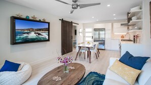 Un salon moderne avec un téléviseur diffusant une scène de plage, un coin repas avec une table et des chaises blanches, et une cuisine avec des armoires blanches et un plan de travail en bois.