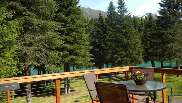 A wooden deck with a table and chairs, overlooking a grassy area with trees and a mountain in the background.