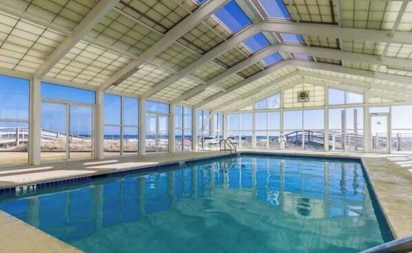 An indoor swimming pool with clear glass walls, a view of the ocean, and a high ceiling with skylights.