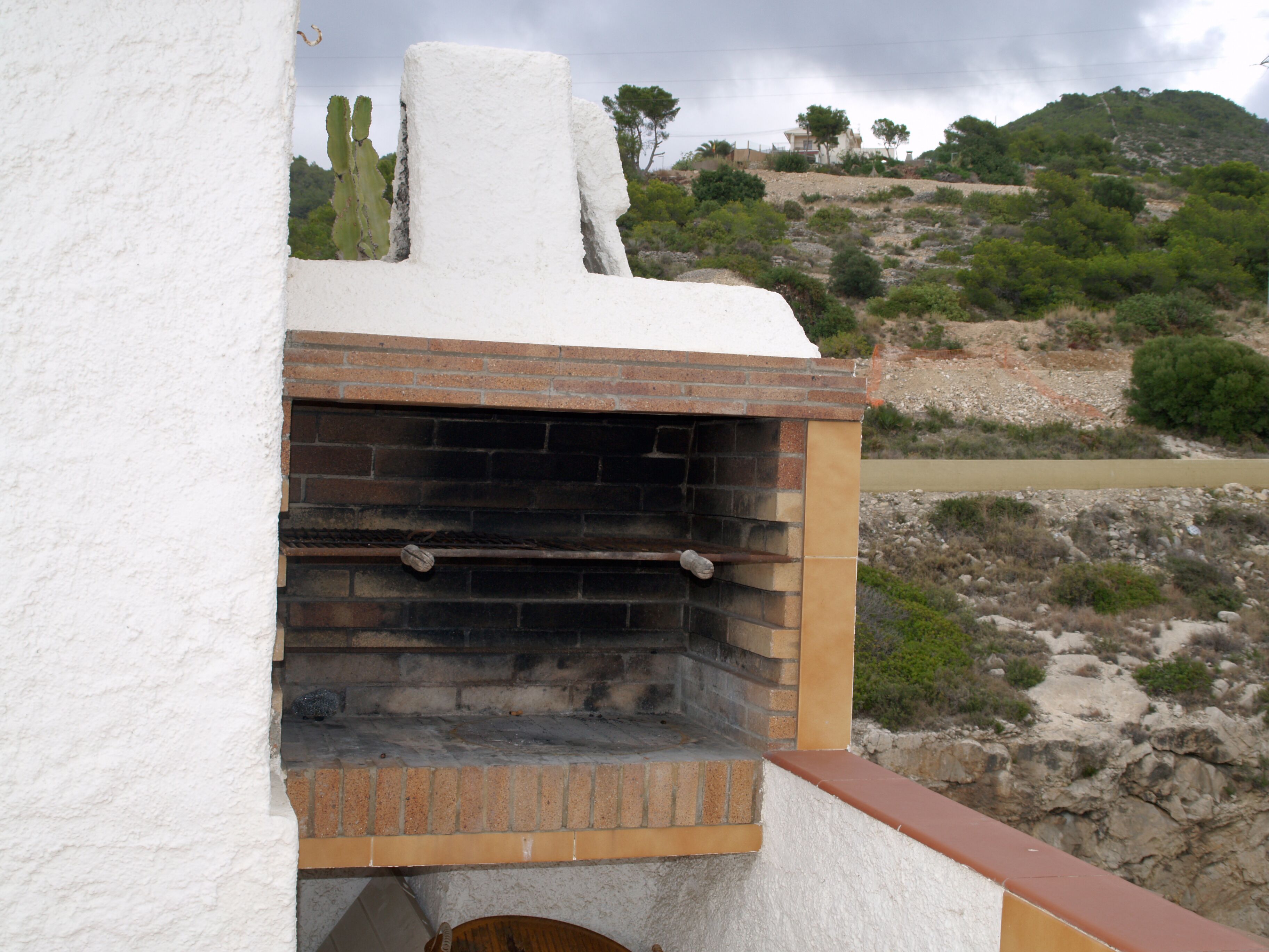 Horno de ladrillo al aire libre adosado a una pared blanca, con vistas a una ladera rocosa y vegetación escasa.