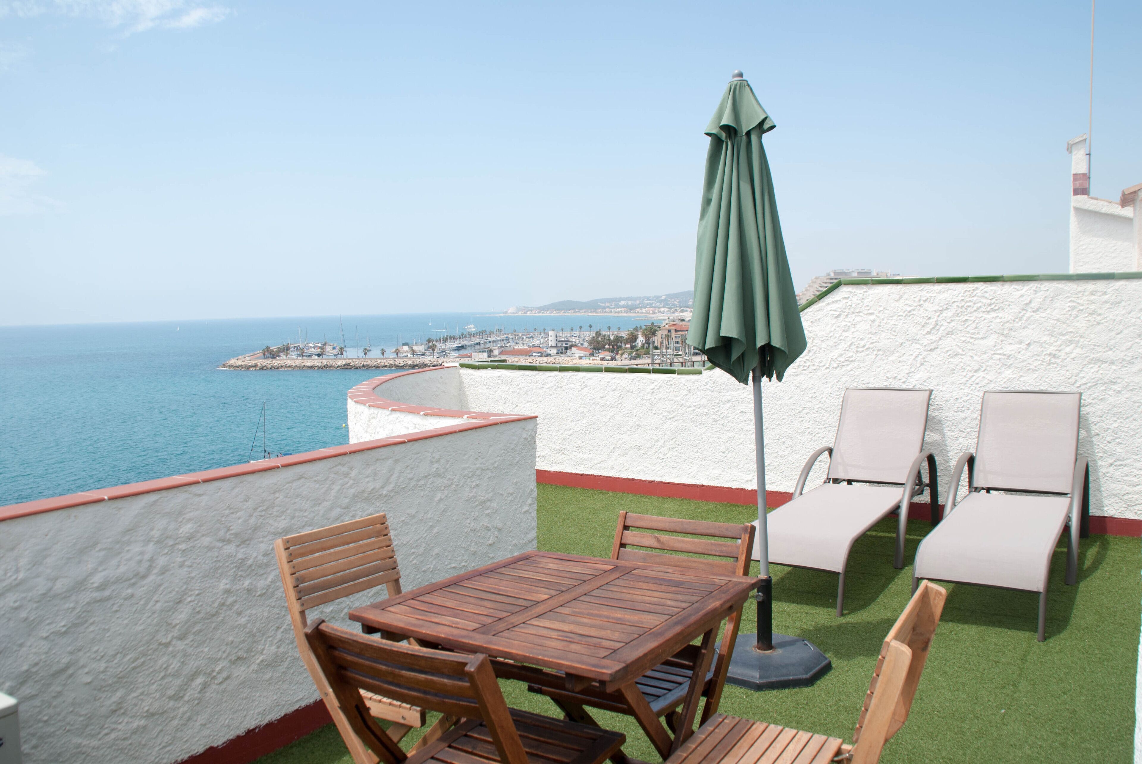 Een terras met een houten tafel en stoelen, een groene parasol en uitzicht op zee en verre gebouwen.