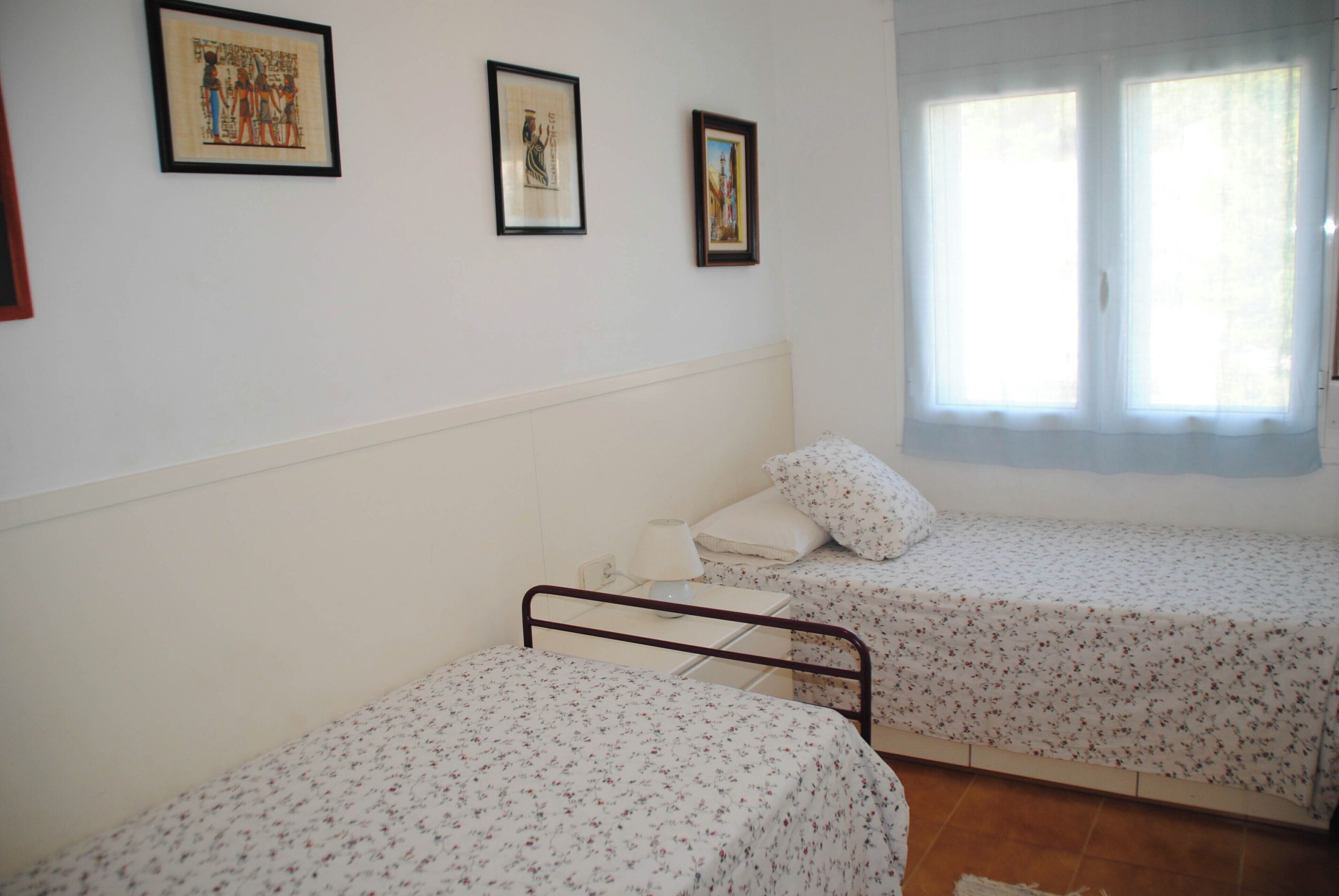 Une chambre avec deux lits simples, une table de chevet avec une lampe et des tableaux encadrés au mur.