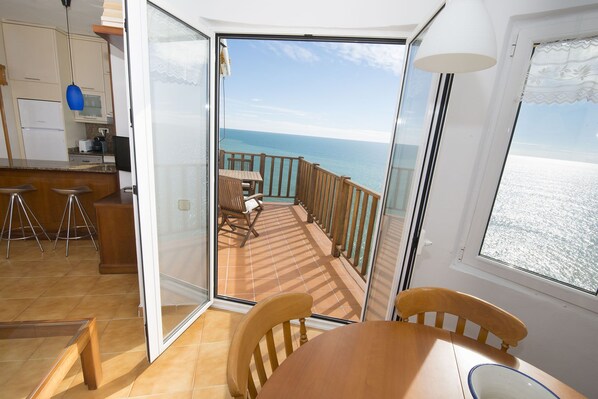 A room with a wooden table and chairs, a kitchen area with a refrigerator and microwave, and a balcony with a view of the sea.