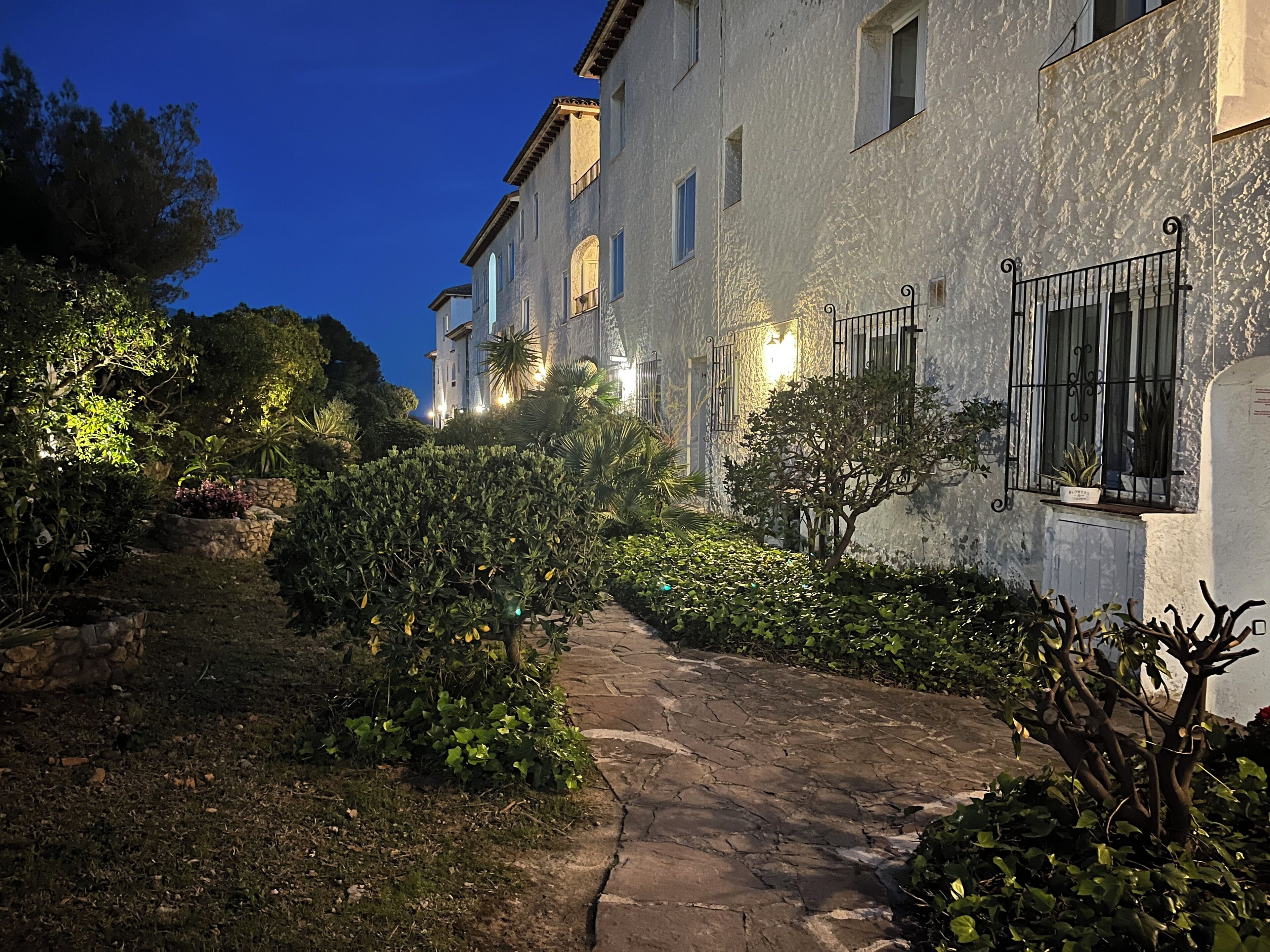 A stone pathway leading to a building with multiple windows and balconies, surrounded by greenery and stone walls.