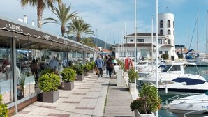 Un port de plaisance avec des bateaux amarrés, une promenade couverte bordée de boutiques et des palmiers.