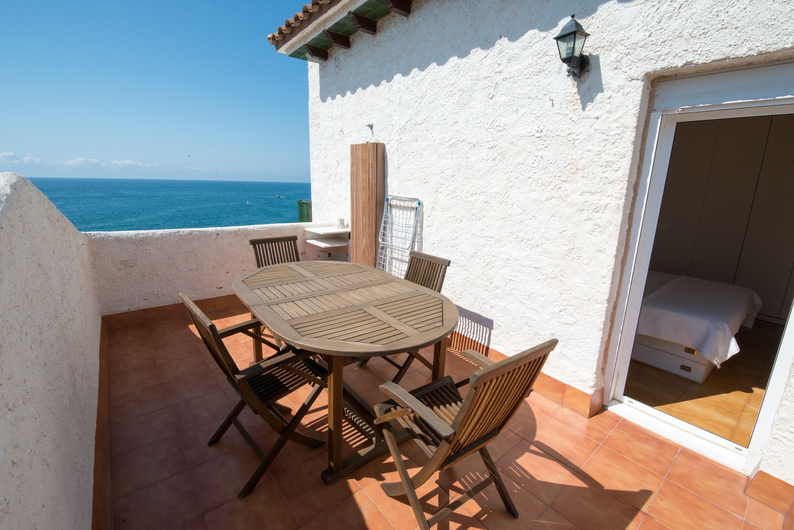 A patio with a round table and chairs, a view of the sea, and a white building with a door.