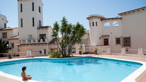 Une piscine où une personne est assise au bord, entourée de palmiers et de bâtiments d’un style architectural méditerranéen.