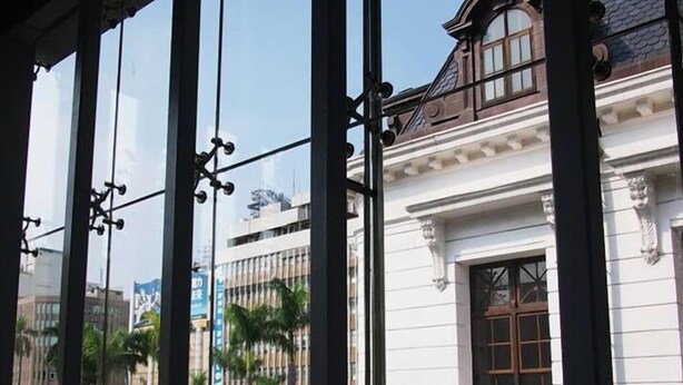 A view through a window with a balcony, featuring a building with classical architecture and a street with cars and palm trees. - A Ace Hotel (Taichung)
