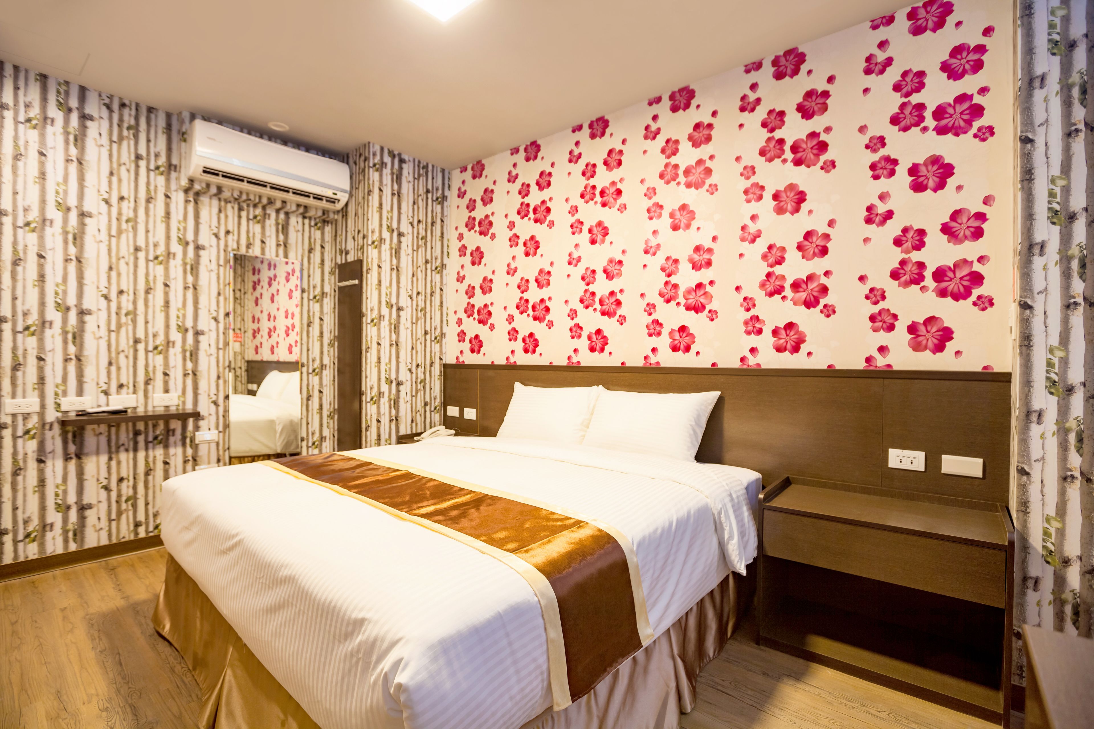 A hotel room with a large bed, a wooden headboard, a bedside table, and a wall with floral wallpaper.