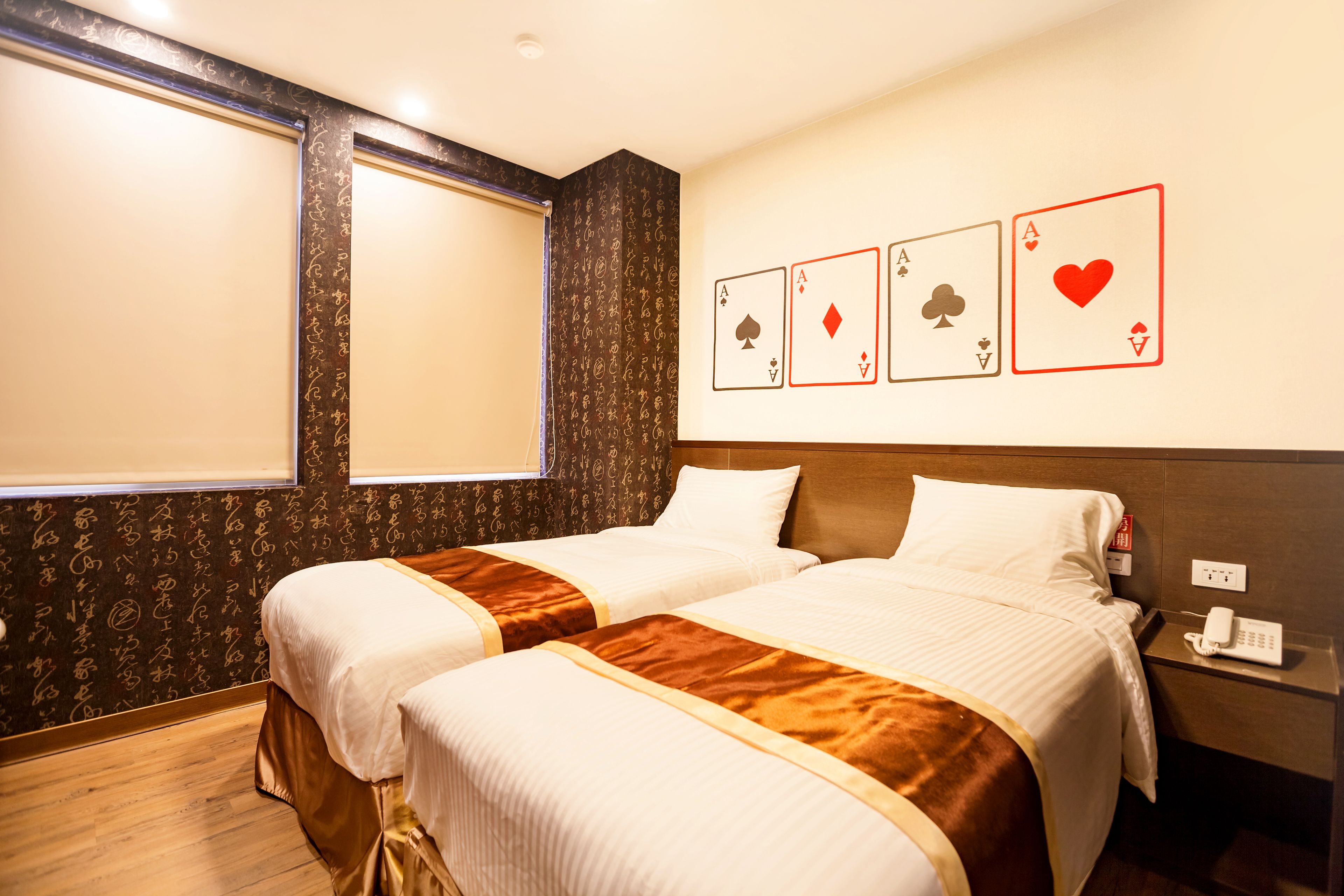 A hotel room with two beds, a wooden floor, a window, and a wall with playing cards.
