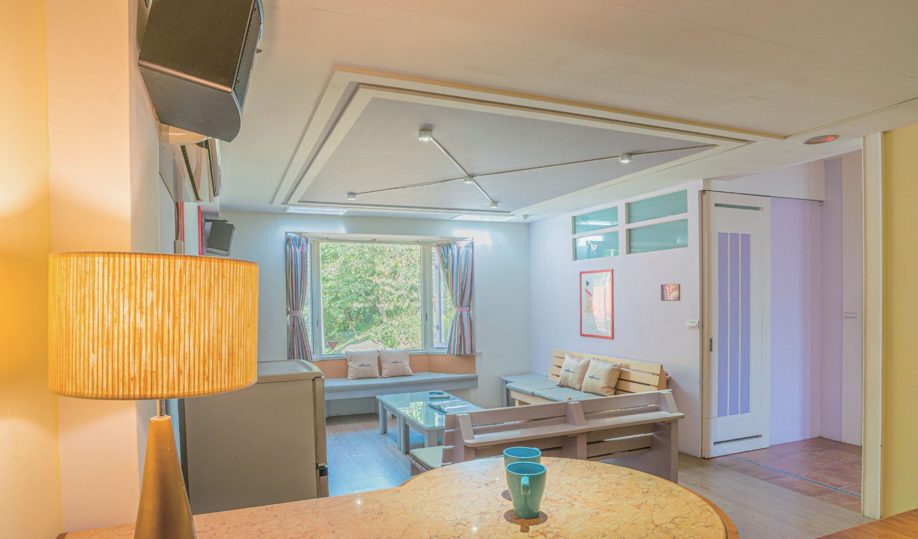 A cozy room with a bed, a small table with a lamp, a bench, and a window with curtains.