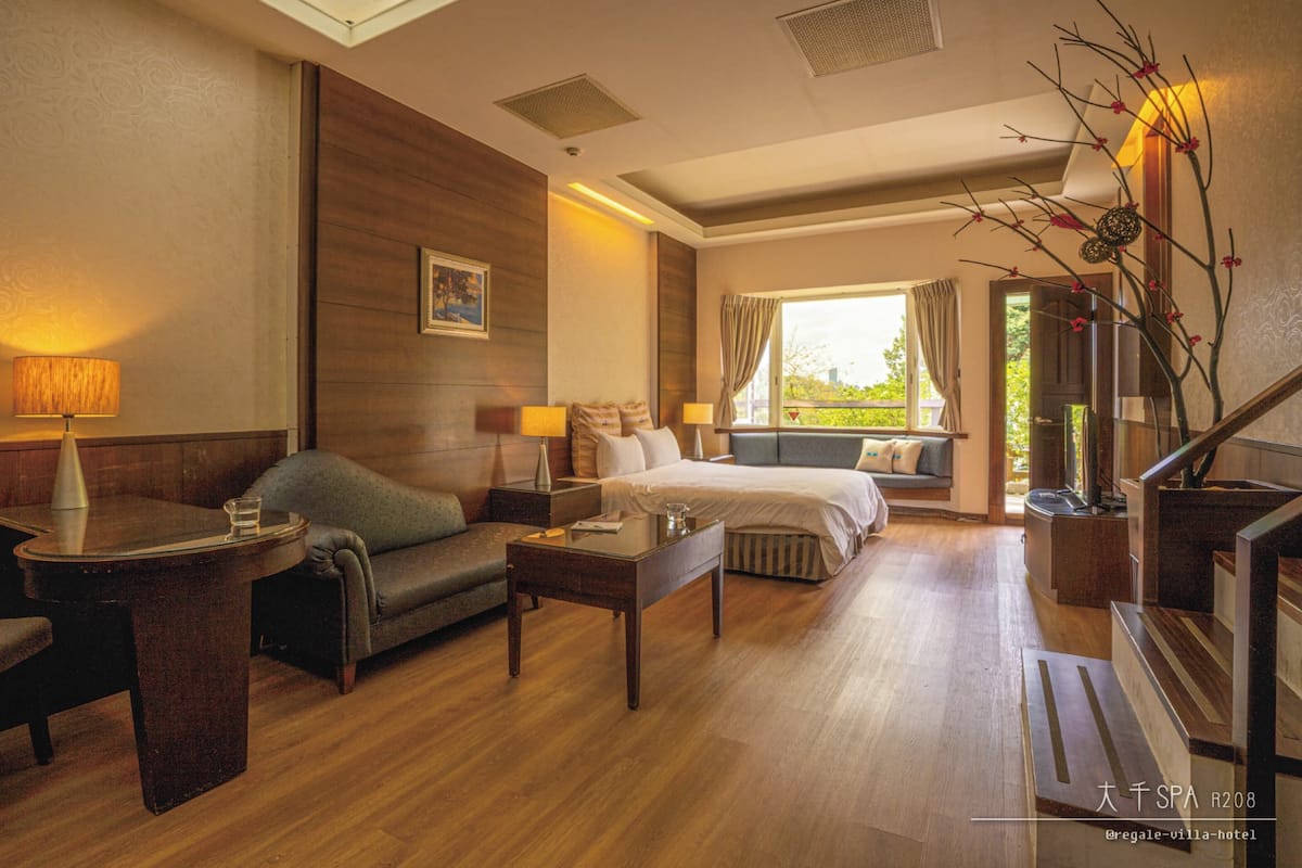 Een hotelkamer met een houten vloer, een bank, een bed, een bureau en een raam met gordijnen.