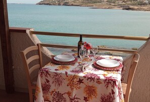 Une table dressée pour deux personnes, avec une nappe à motifs floraux, une bouteille et des verres, donnant sur une vue sur la côte.