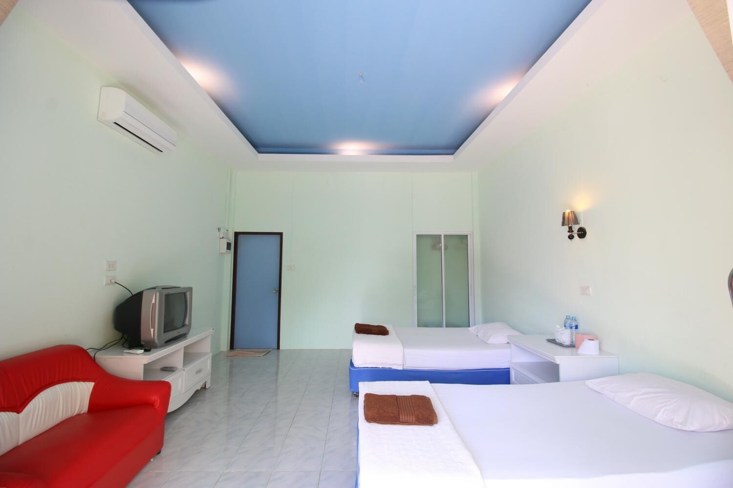 Quarto de hotel com televisão, sofá vermelho, duas camas, guarda-roupa e casa de banho com porta de vidro.
