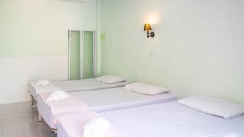 A room with three single beds, each with a pillow and a folded towel. There is a light switch and a wall-mounted light fixture on the wall.