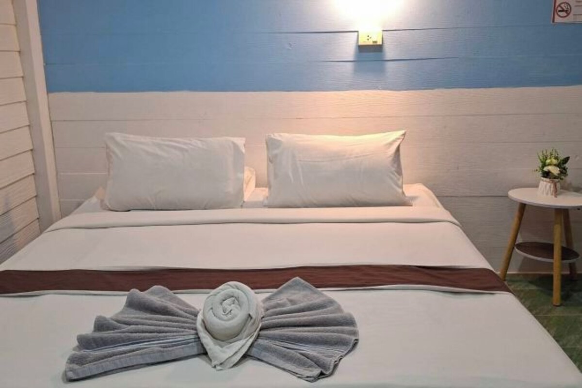 A neatly made bed with white linens and a brown accent, a small round table with a plant, and a wall-mounted light fixture.
