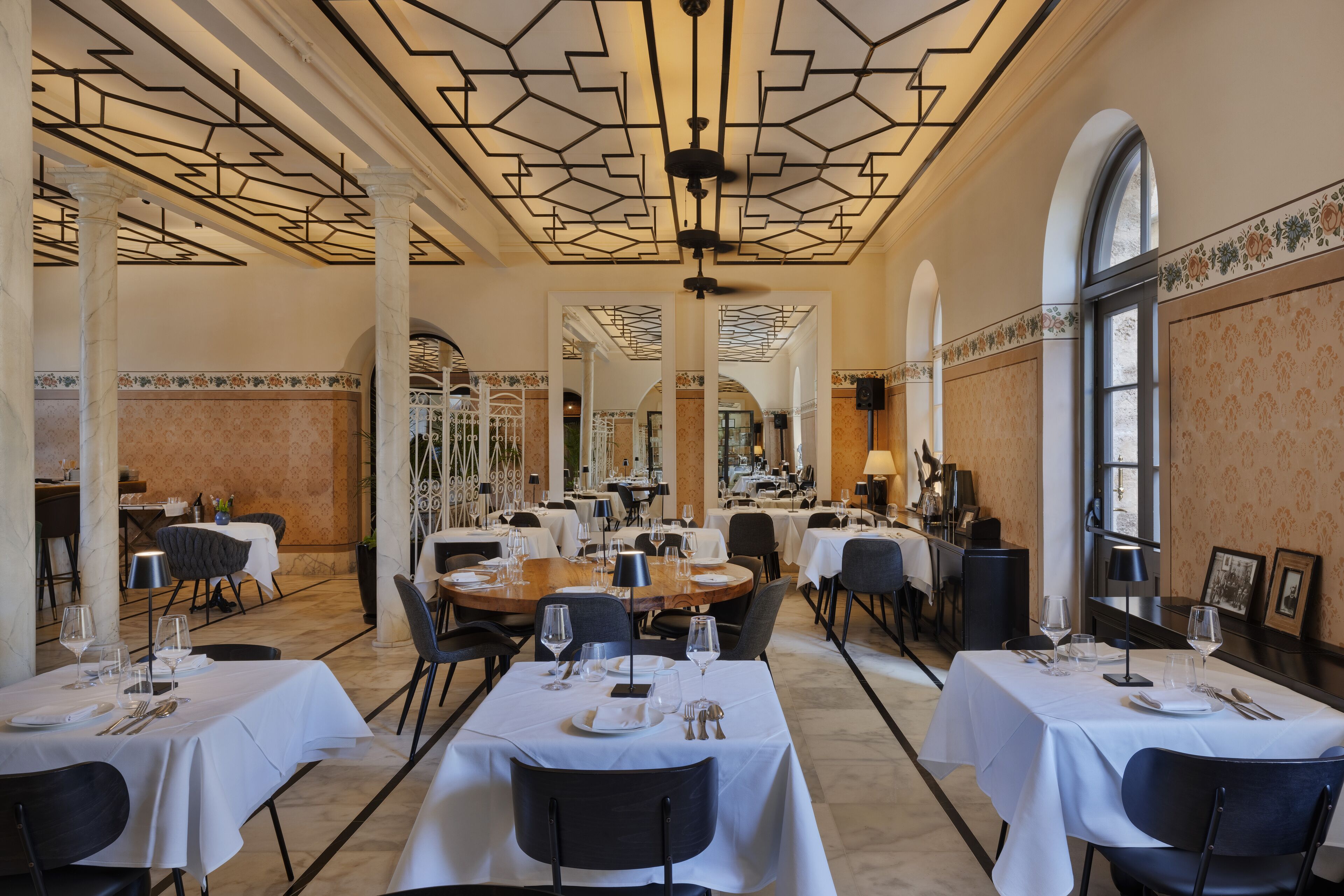 Un vaste espace dédié à la salle à manger, avec plusieurs tables dressées, un plafond au design géométrique et de grandes fenêtres.