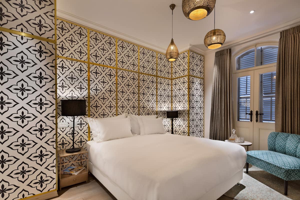Une chambre d’hôtel avec un grand lit, deux tables de chevet avec des lampes, une petite table ronde, une chaise à motifs bleus et une fenêtre avec des rideaux.