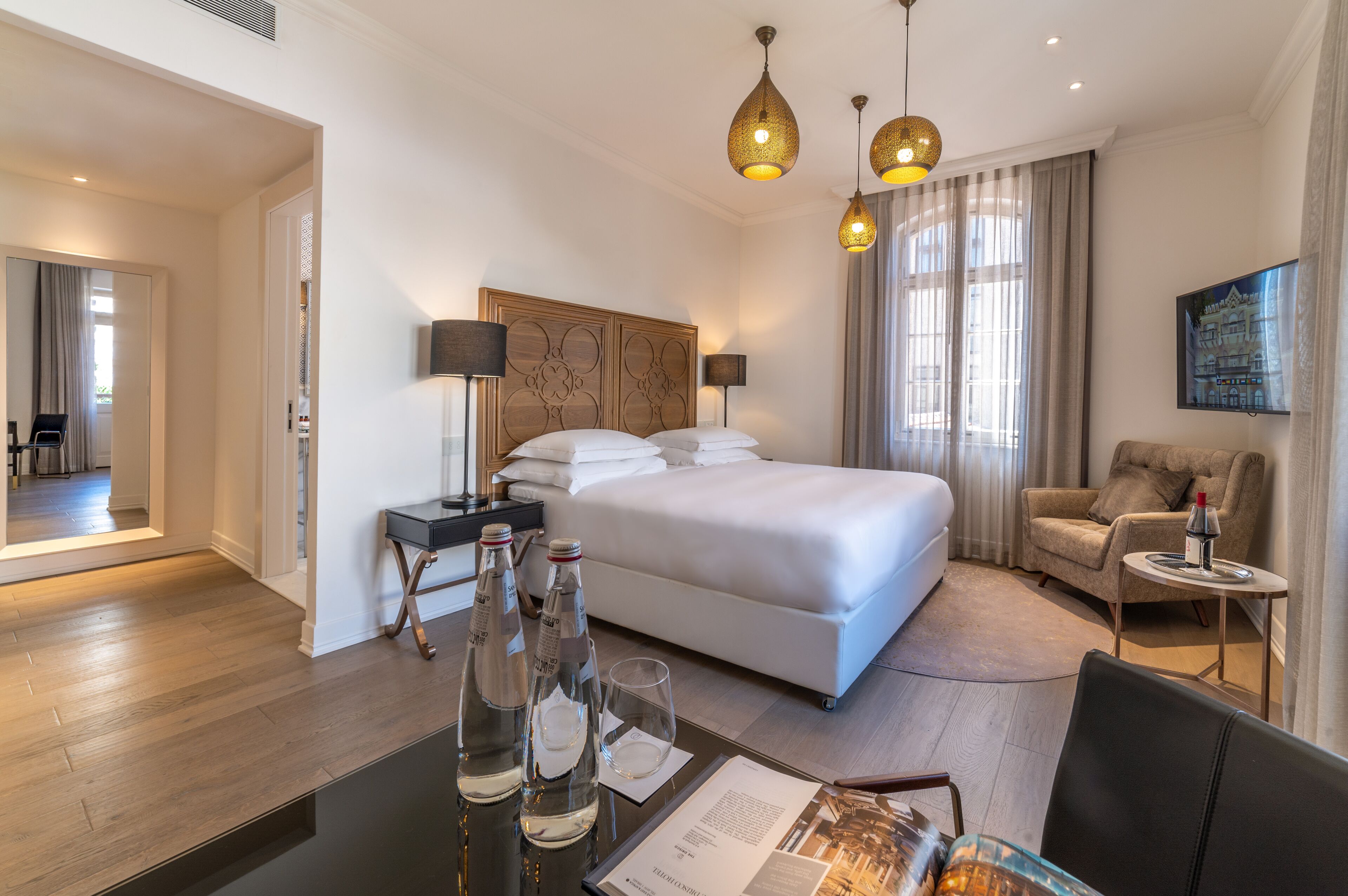 Ein Hotelzimmer mit einem großen Bett, Nachttischlampen, einem Schreibtisch mit Stuhl, einem kleinen Tisch mit Lampe und Blick auf die Stadt durch das Fenster.