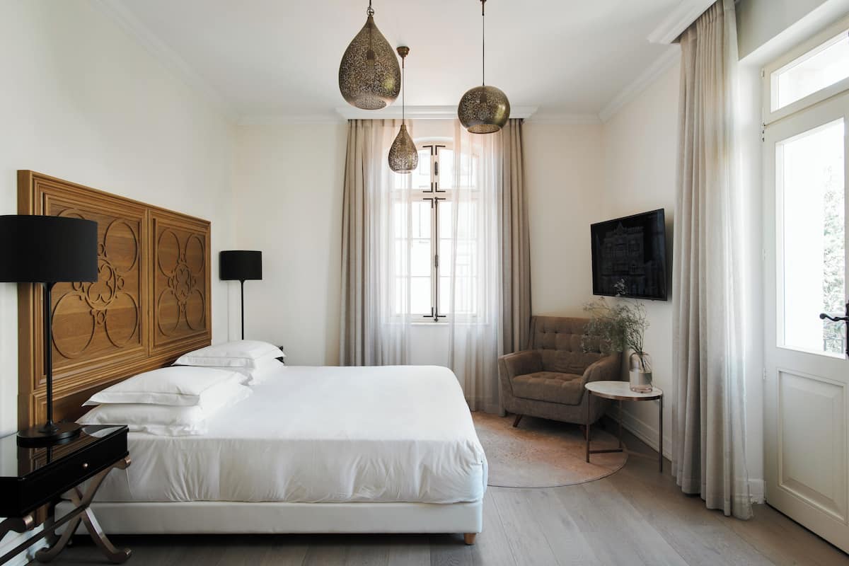 Une chambre d’hôtel avec un grand lit, une tête de lit en bois, un téléviseur fixé au mur, une petite table avec une plante et une fenêtre avec des rideaux.