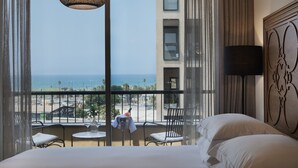 A hotel room with a large window overlooking a beach and ocean, a bed, a small table with a bottle, and a chair. - The Drisco Hotel Tel Aviv - Relais & Châteaux (Tel Aviv)