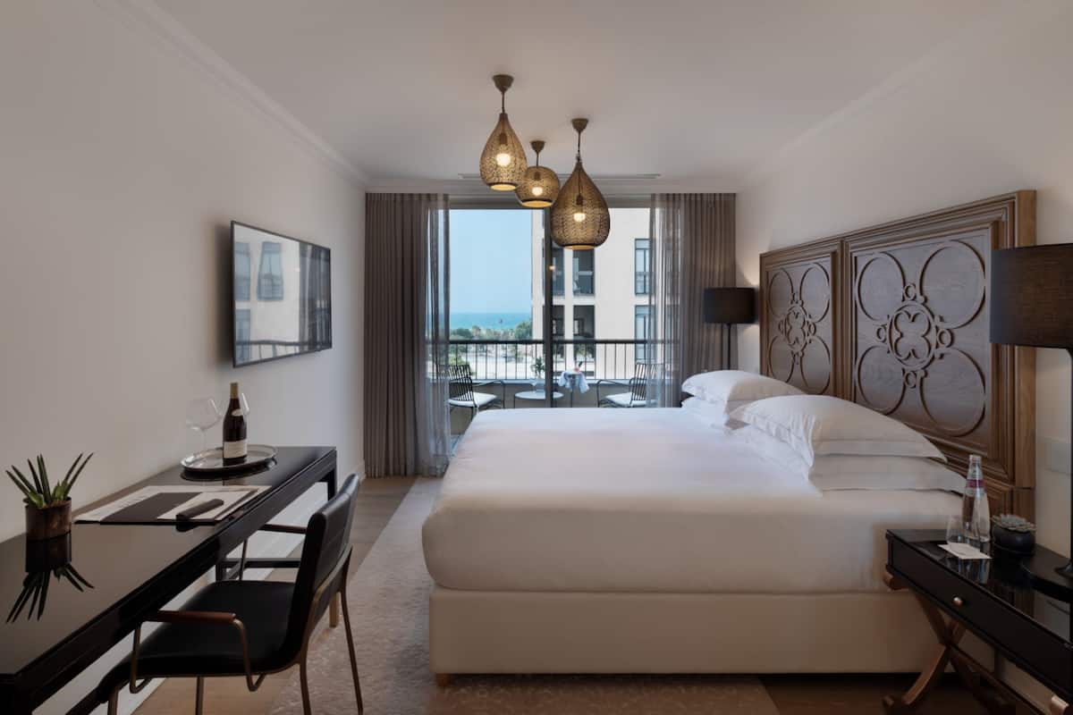 Une chambre d’hôtel avec un grand lit, un bureau avec une chaise, une petite table avec une chaise, et un balcon offrant une vue sur la ville.