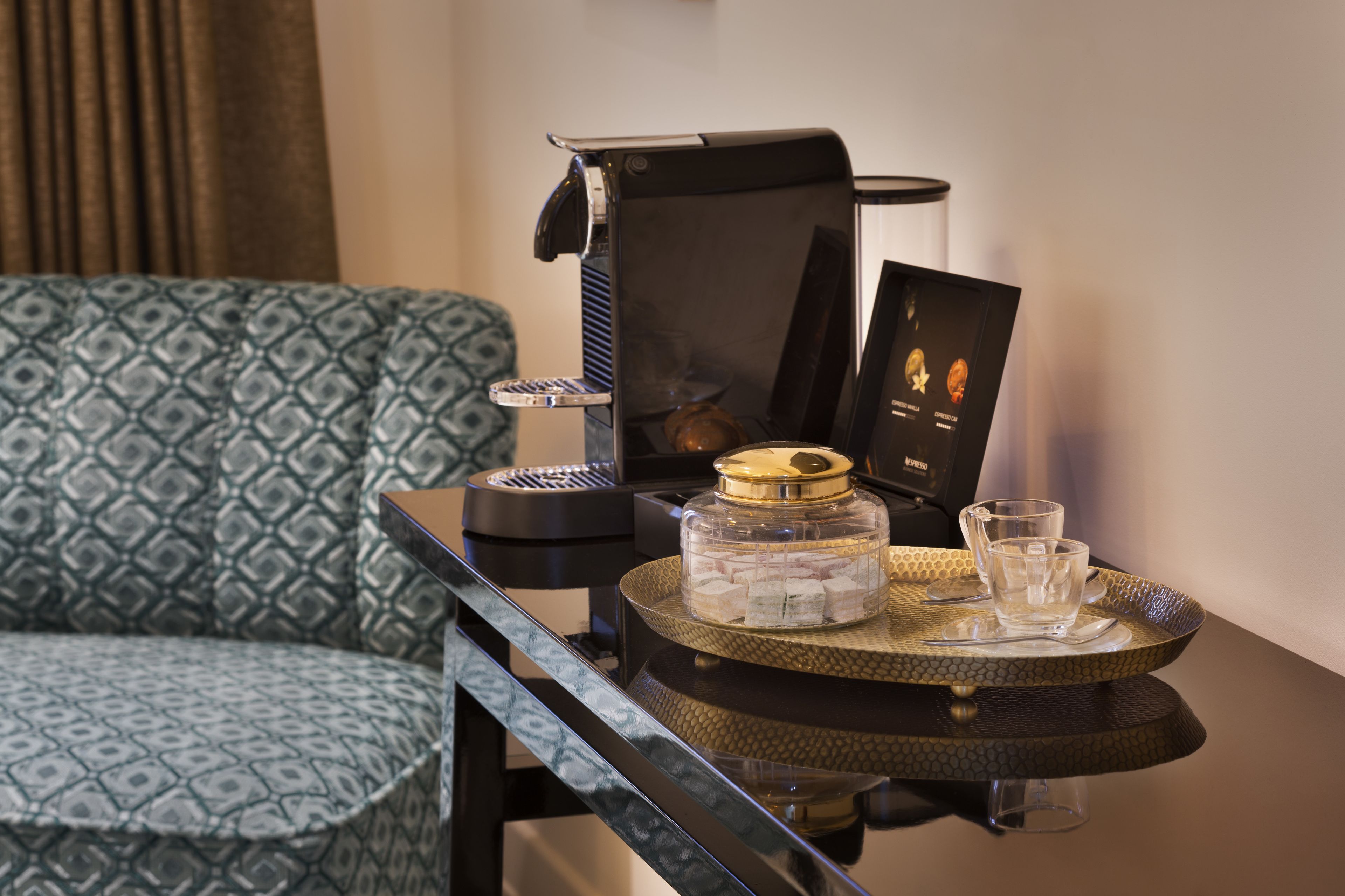 A room with a patterned sofa, a small round table with a tray, a coffee machine, and a decorative item.