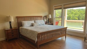 A bedroom with a wooden bed, two bedside tables with lamps, a large window with a view of greenery, and a balcony. - Beautiful lake front house, pool table, and fire pit, kayaks,Fall price discount (Hiawassee)